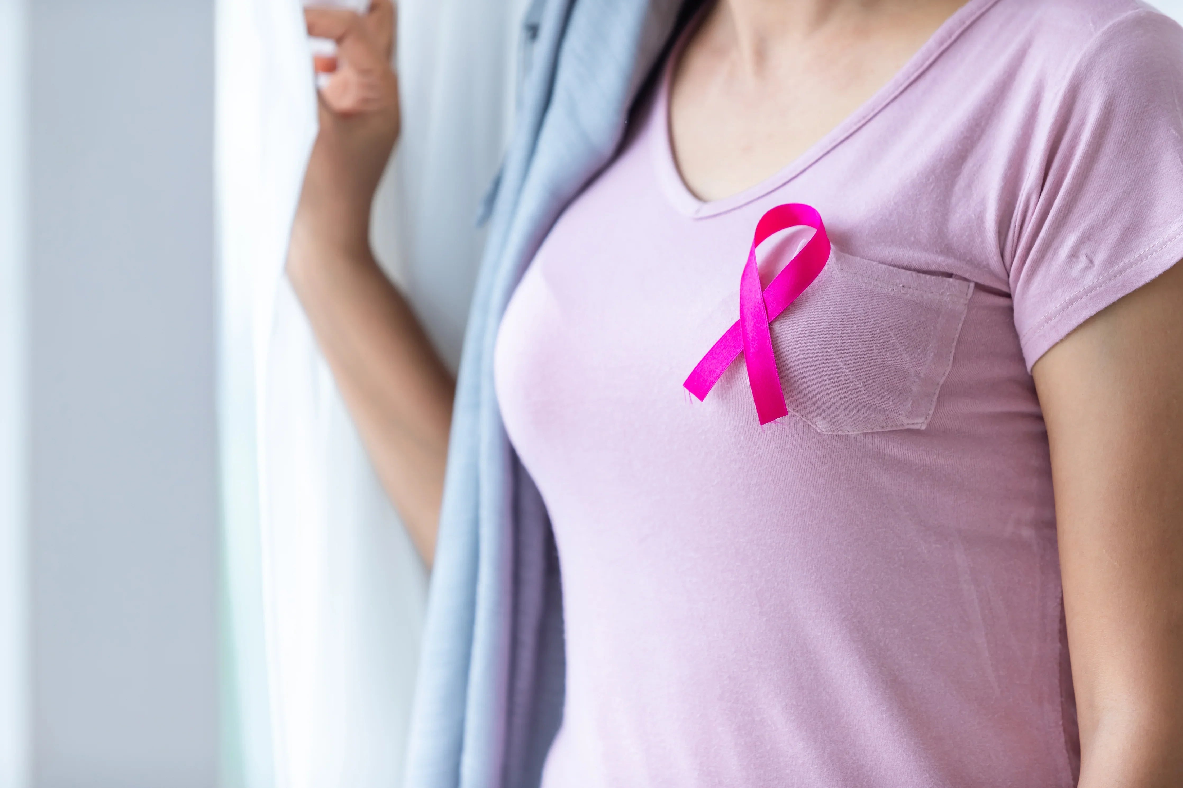 Woman in soft pink shirt with pink ribbon, breast health awareness, near window