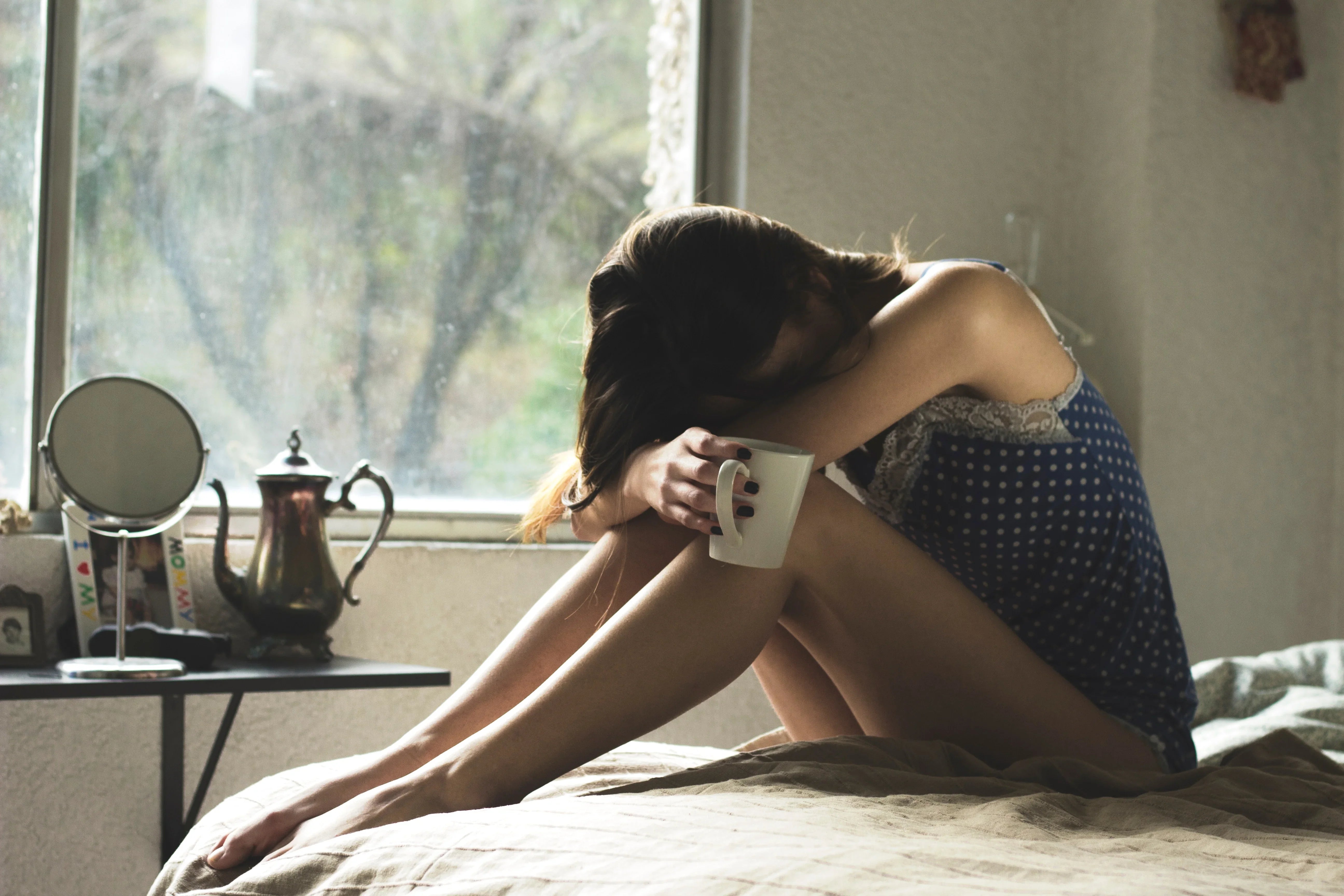 Woman in blue polka dot hypoallergenic nightwear sitting on bed with a mug, near window