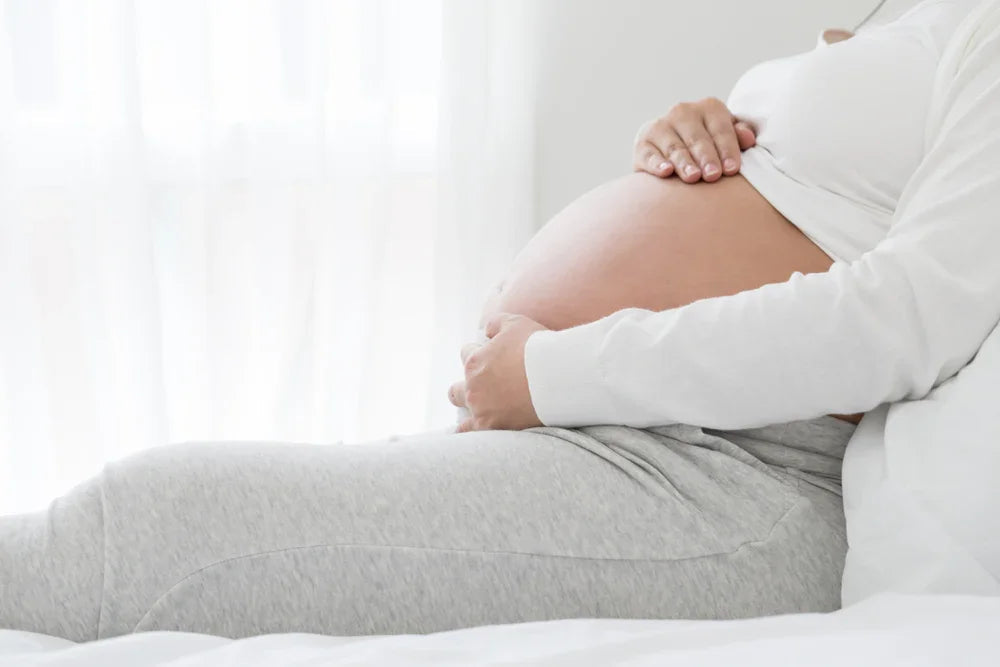 Pregnant woman in white organic cotton loungewear holding belly, sitting on bed
