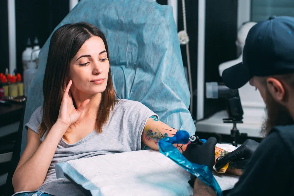 Woman in a gray allergy-free shirt getting a colorful arm tattoo in a studio setting