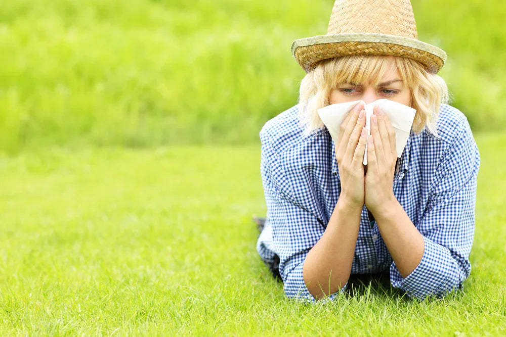 Woman with allergies sneezing in grassy field, wearing a blue checked shirt and straw hat