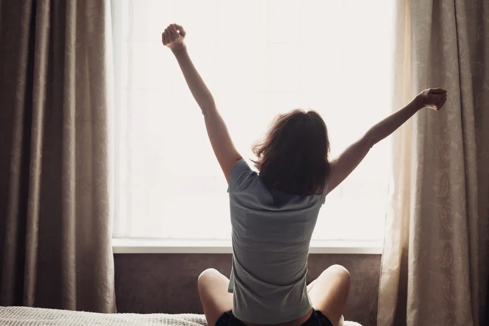 Person in soft cotton sleepwear stretching on bed in morning sunlight by window