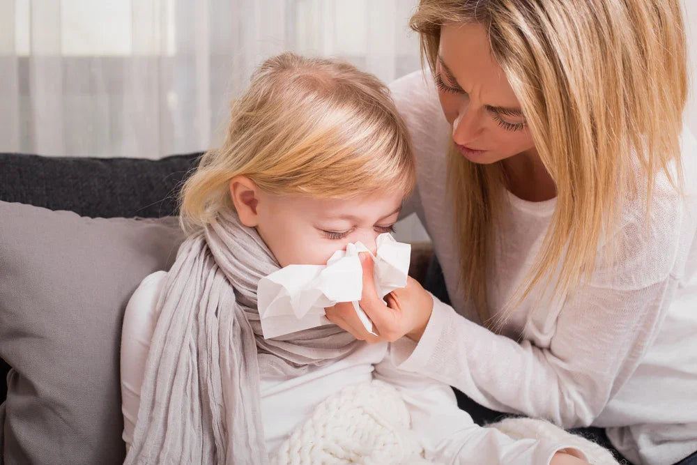Mother helping child with allergies by wiping nose, both wearing soft cotton-like clothing on couch.