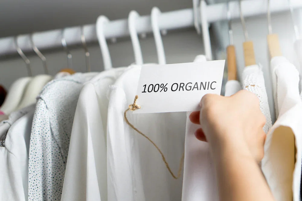 Hand holding 100% organic tag near white cotton shirts on hangers for sensitive skin