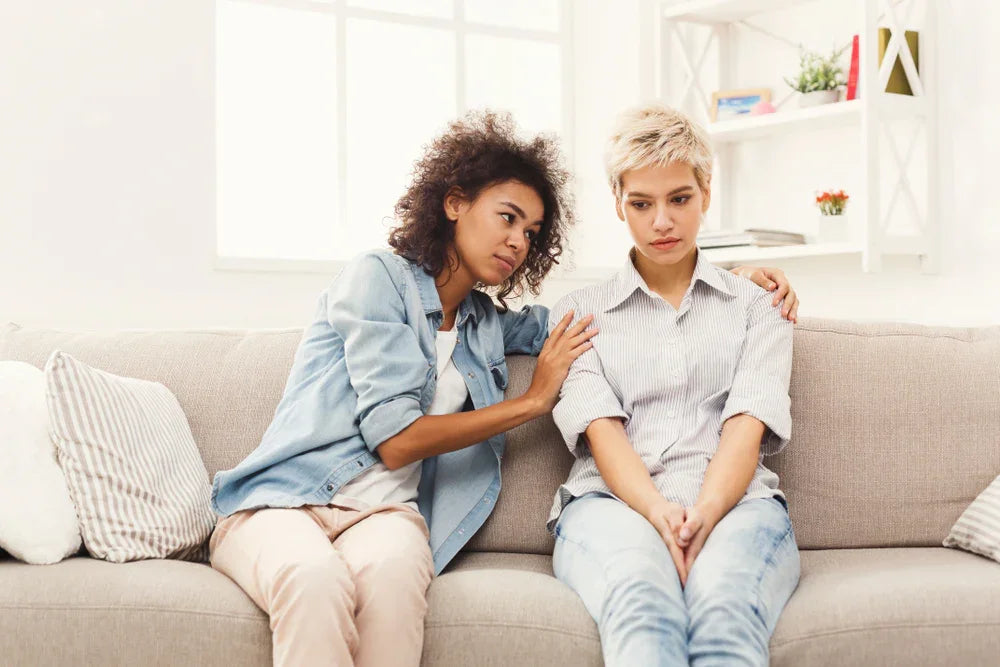 Two women in casual cotton clothing sit on a couch, one comforting the other, bright living room.