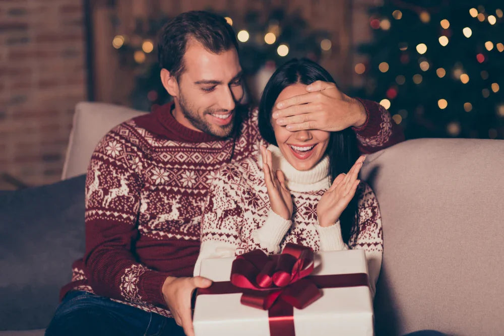 Happy couple in matching winter sweaters exchanging a holiday gift on a cozy sofa