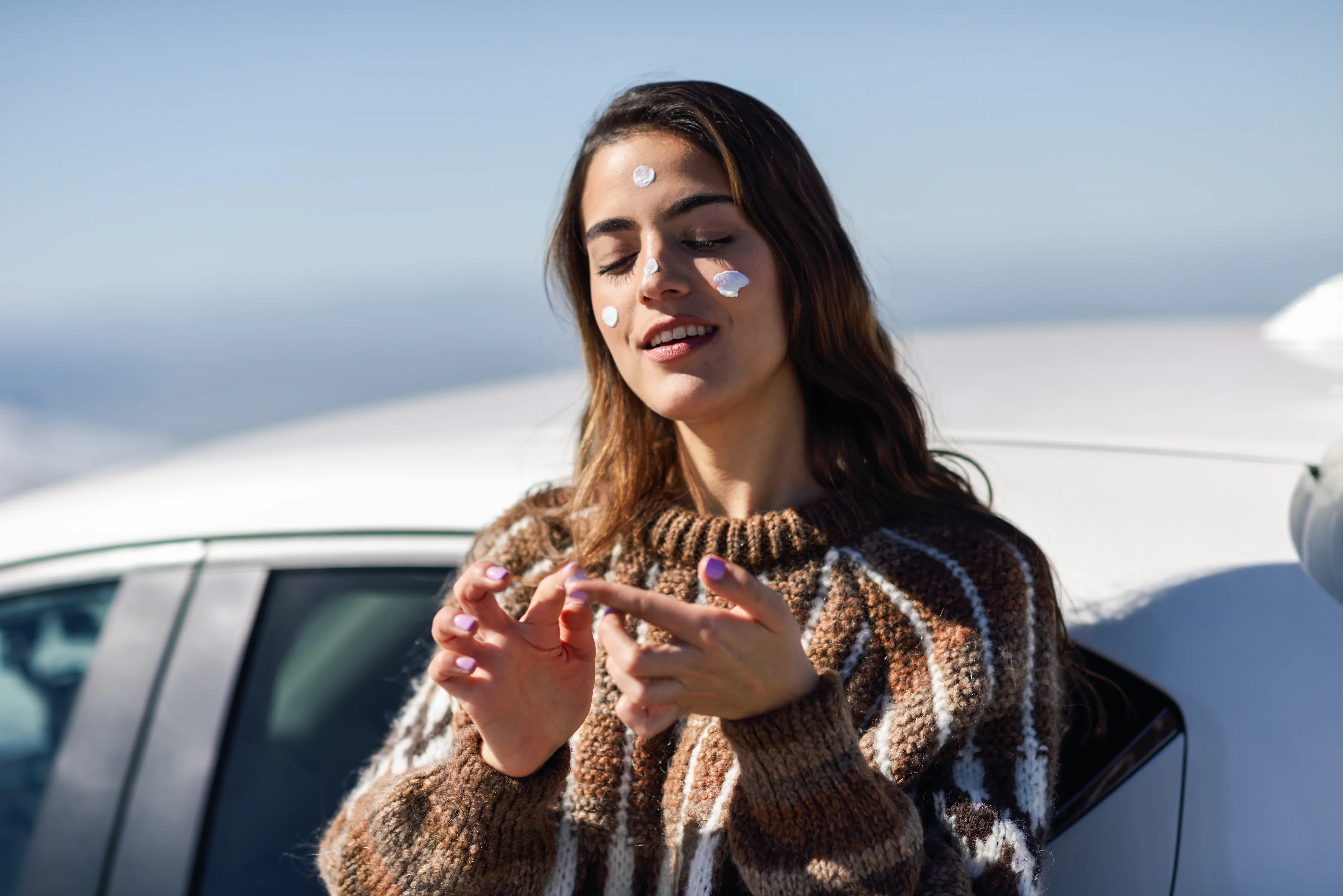Woman with sensitive skin applies sunscreen wearing a knit sweater outdoors by a car