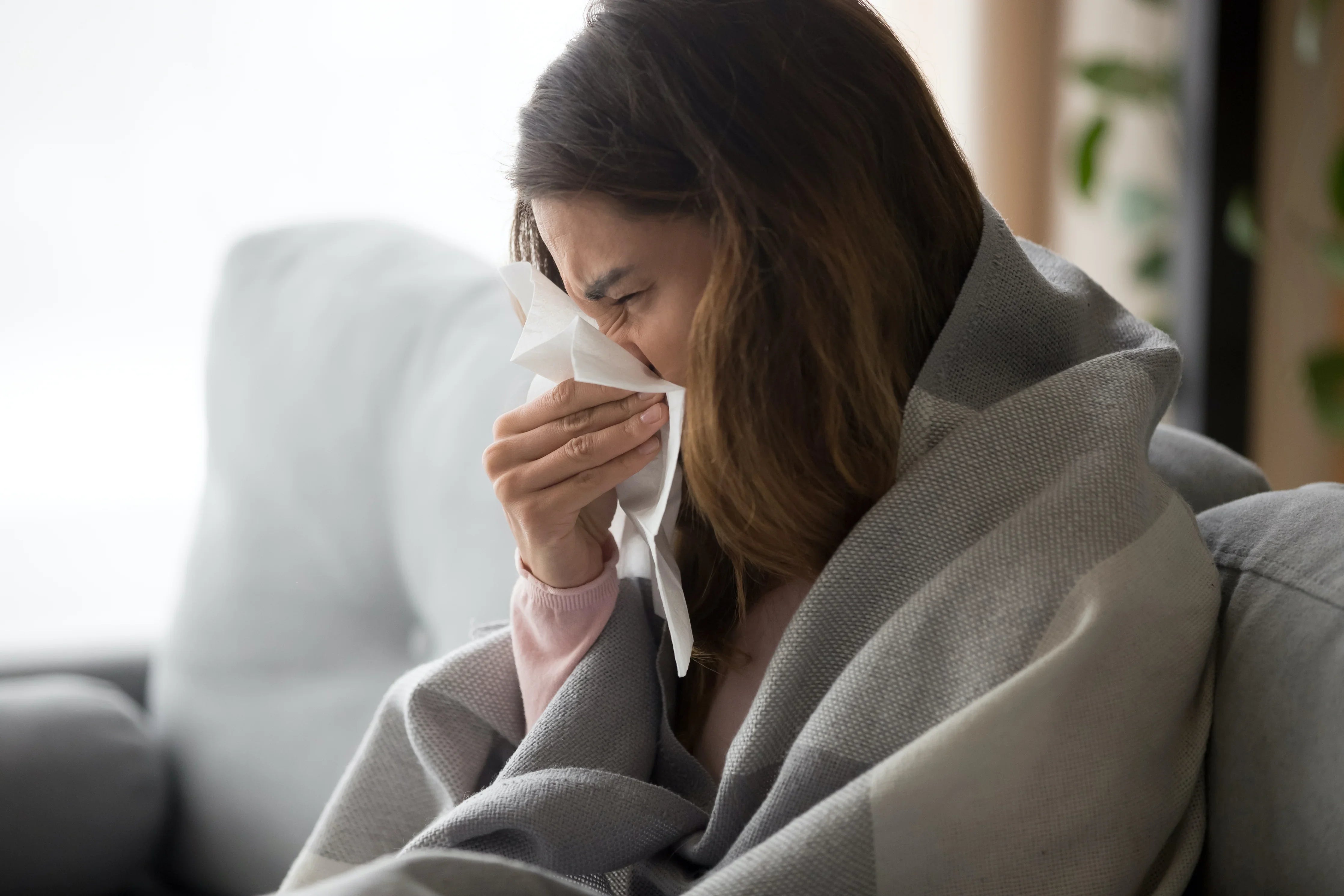 Woman with sensitive skin sneezing under a cozy blanket in a bright living room