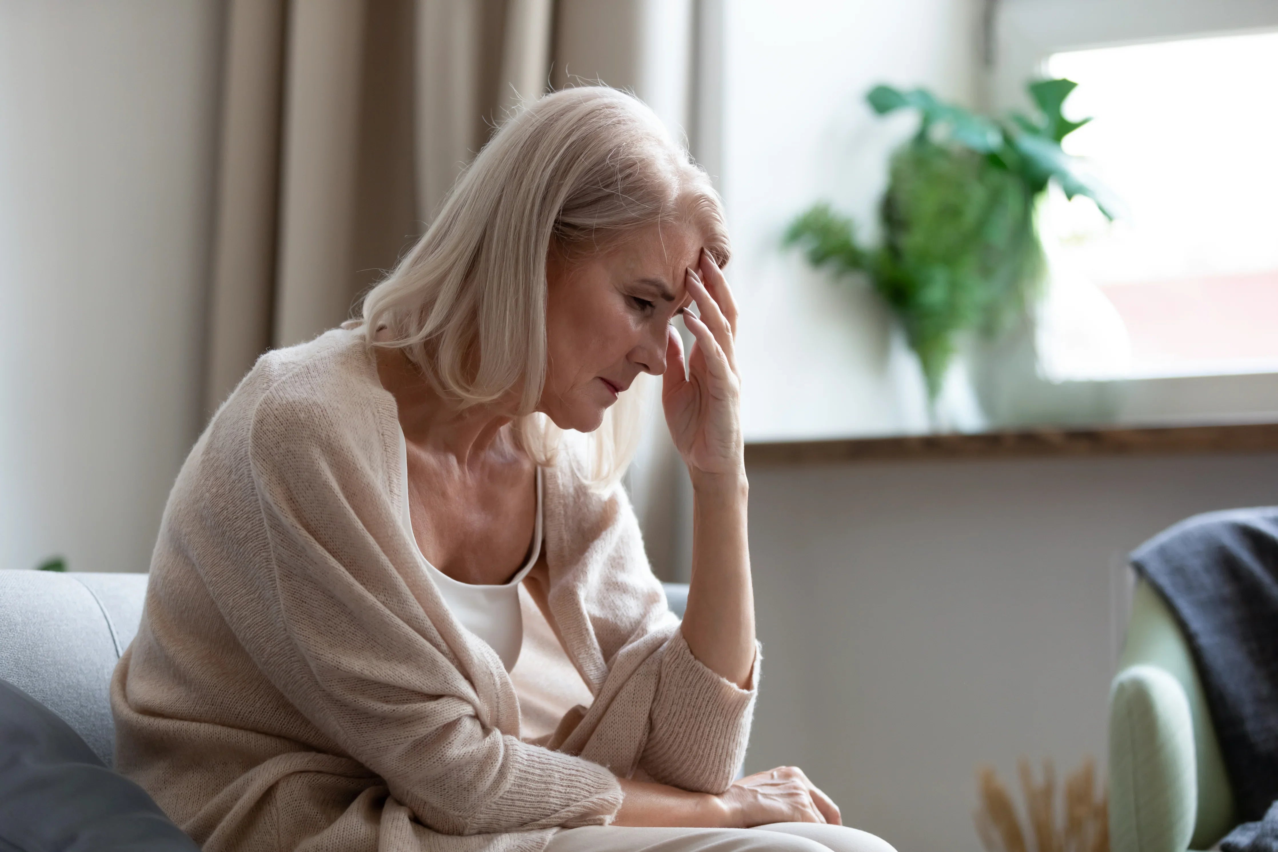 Senior woman with gray hair in soft clothing sitting indoors, looking thoughtful and stressed