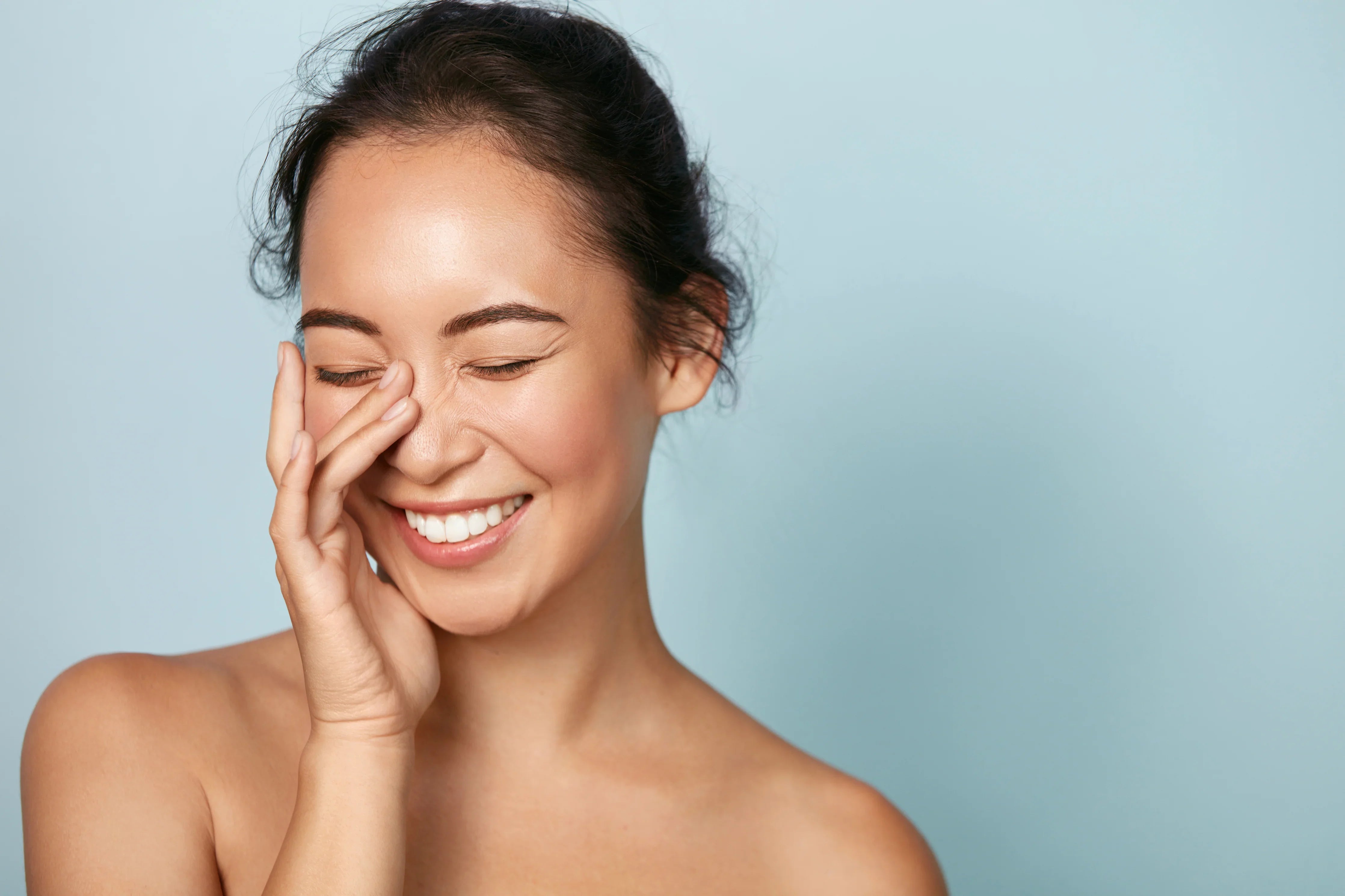 Smiling woman with clear, sensitive skin gently touching her face against a light blue background