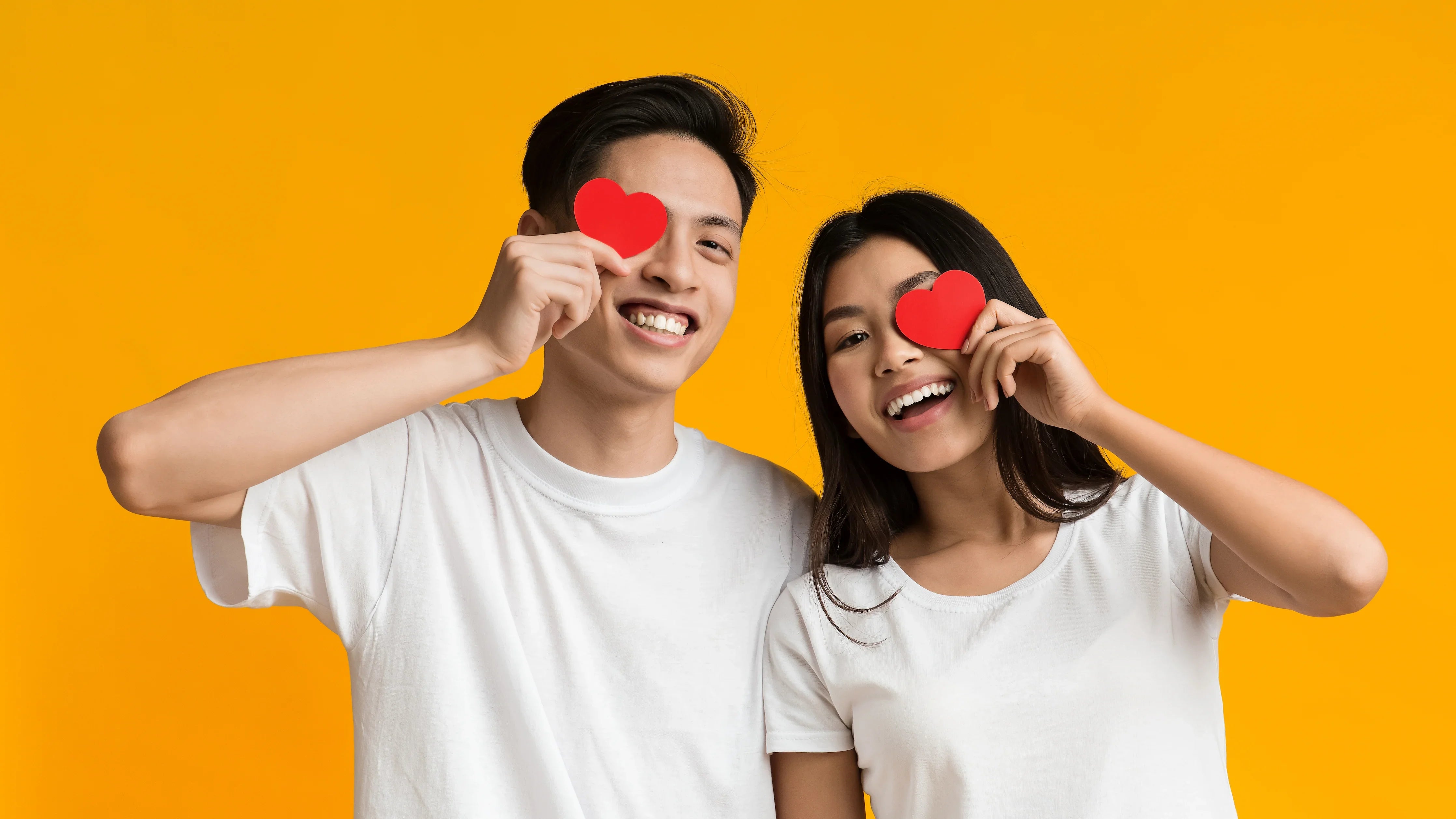 Smiling couple in white cotton t-shirts holding red paper hearts, orange background