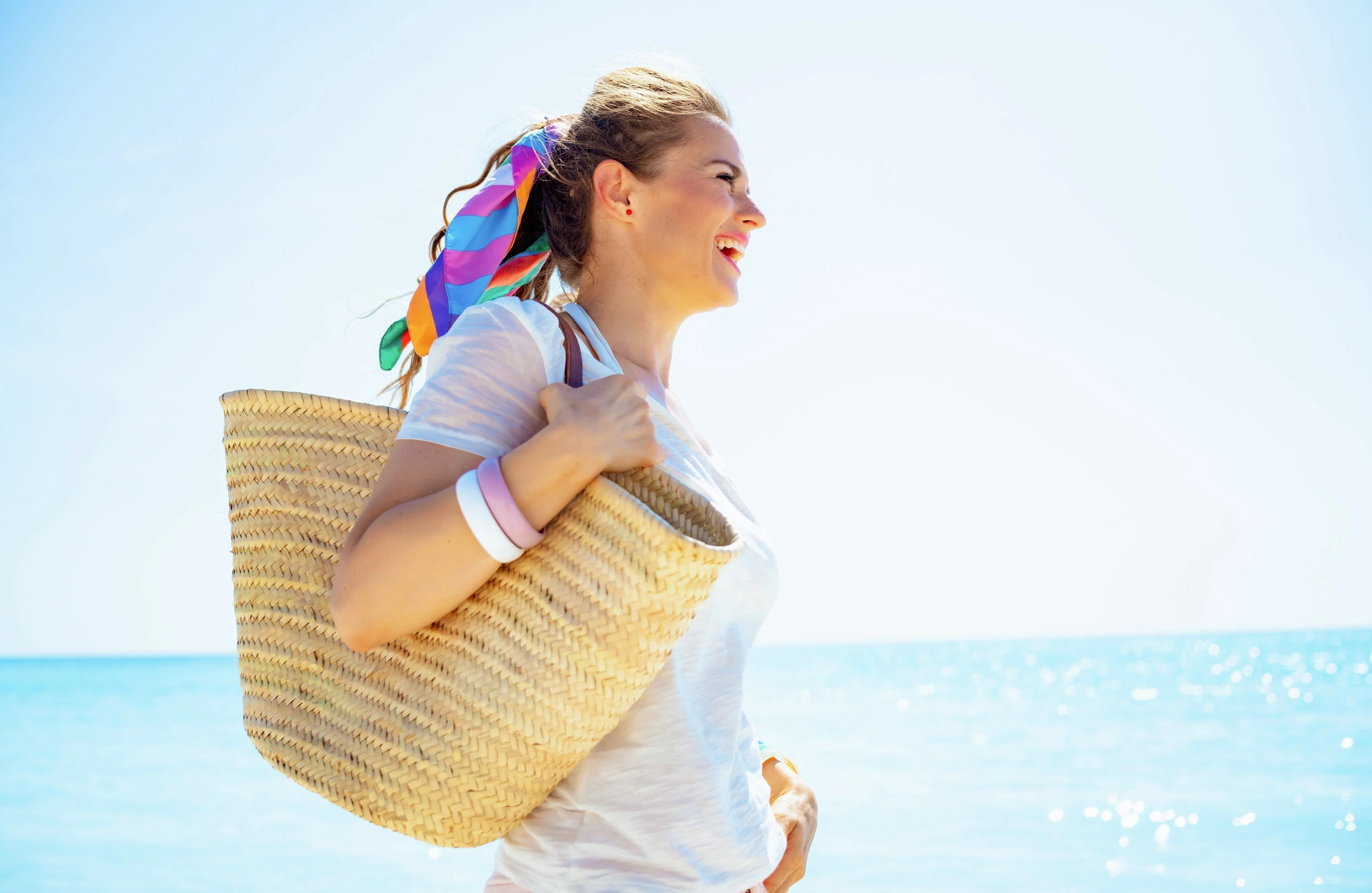 Smiling woman in a white hypoallergenic t-shirt and straw bag on a sunny beach