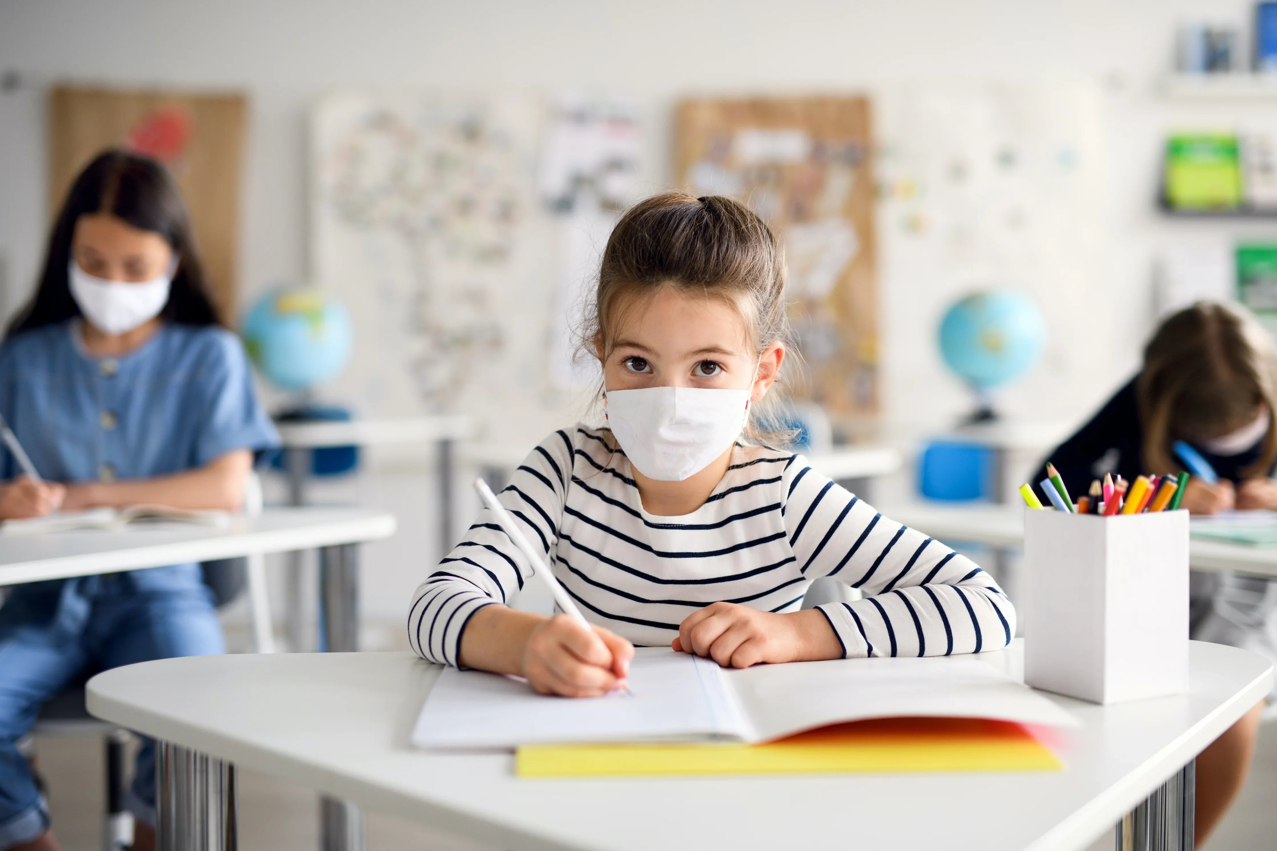 Girl in striped shirt wearing a white face mask writing at desk in a classroom setting