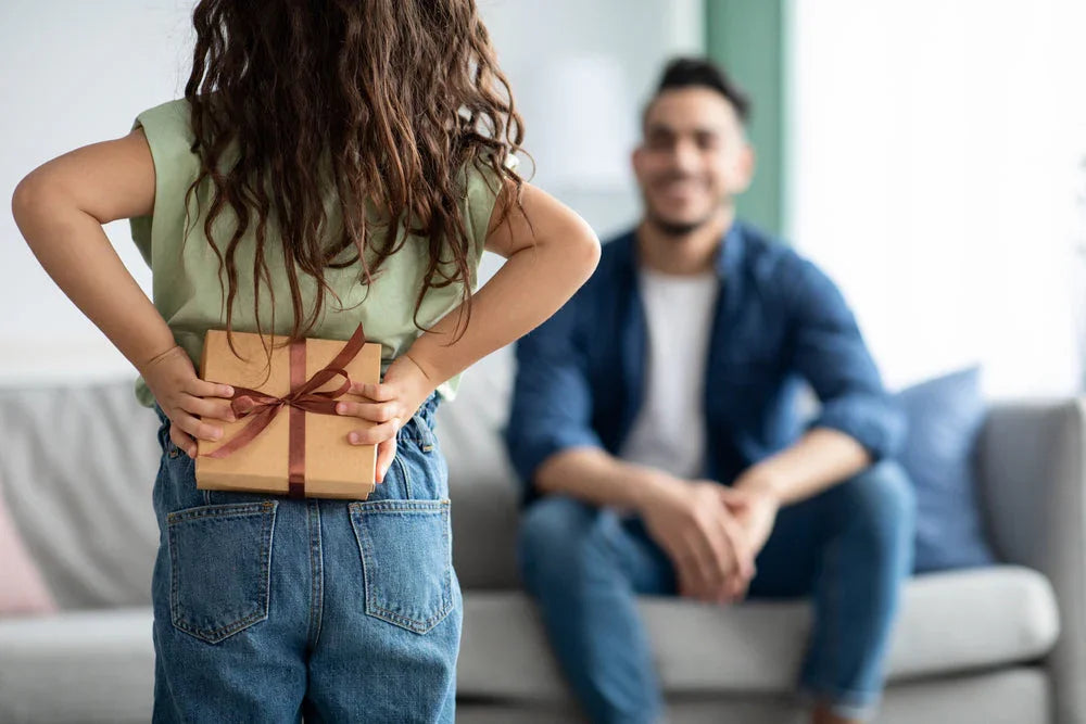 Child in casual cotton clothing hiding gift behind back, adult sitting on sofa in background