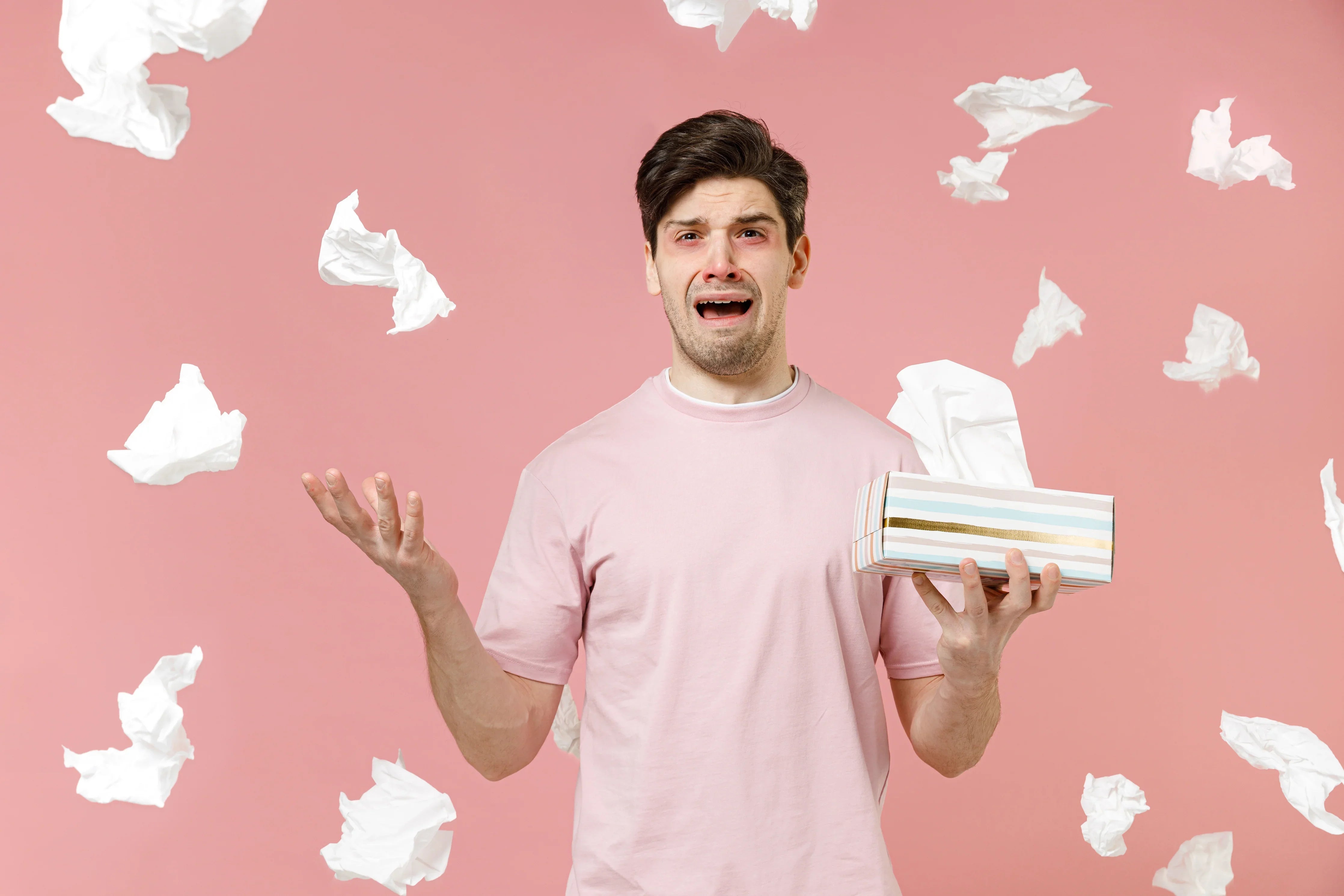 Man with allergies holding tissues, wearing pink T-shirt, surrounded by tissues on pink background