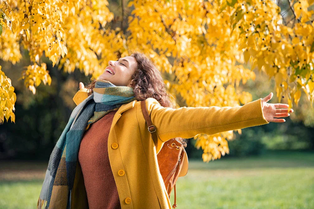 Woman wearing fall organic cotton clothing, arms outstretched in a park with yellow autumn leaves.