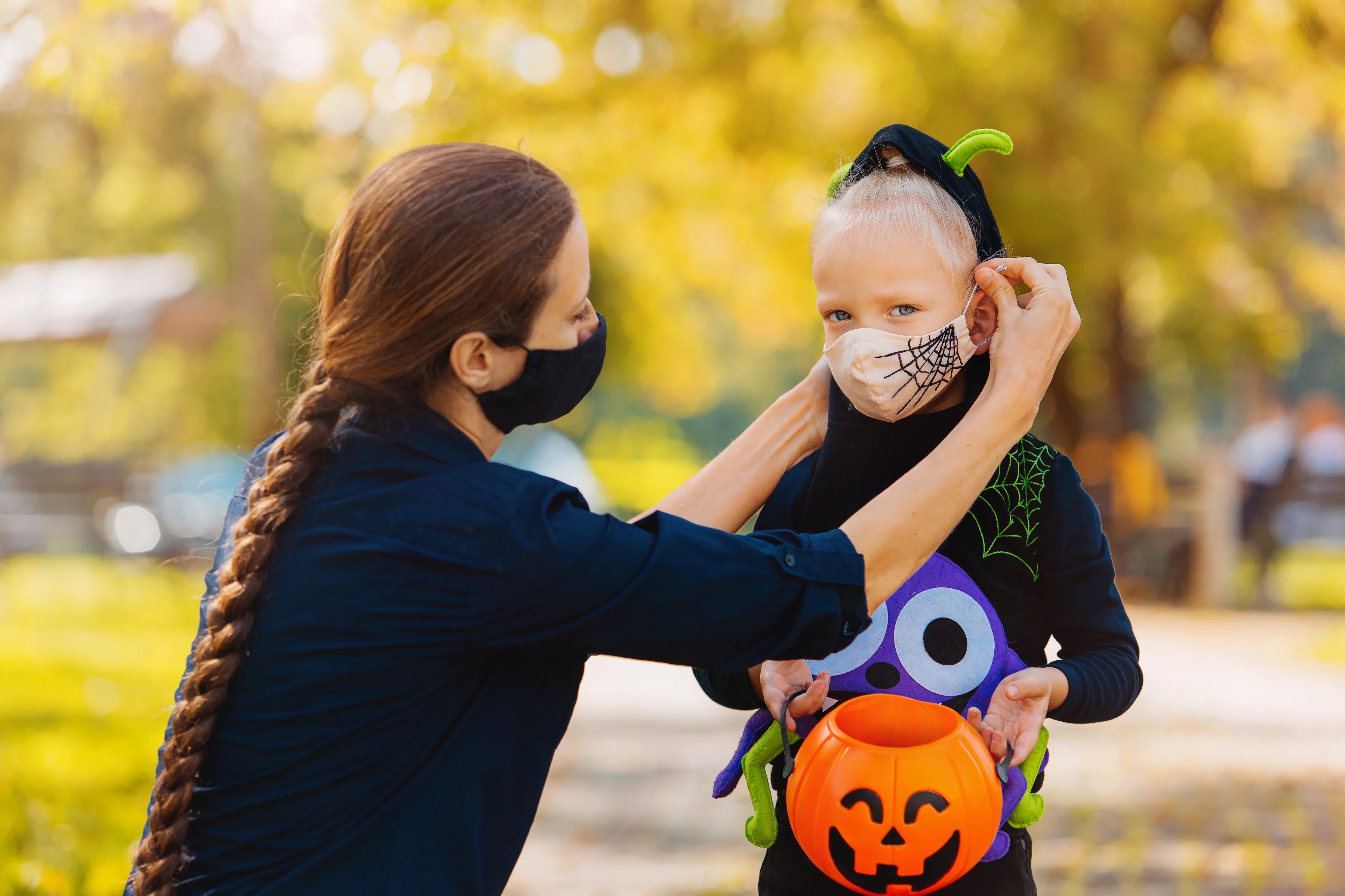 Woman helping child in Halloween costume and face mask with pumpkin bucket outdoors