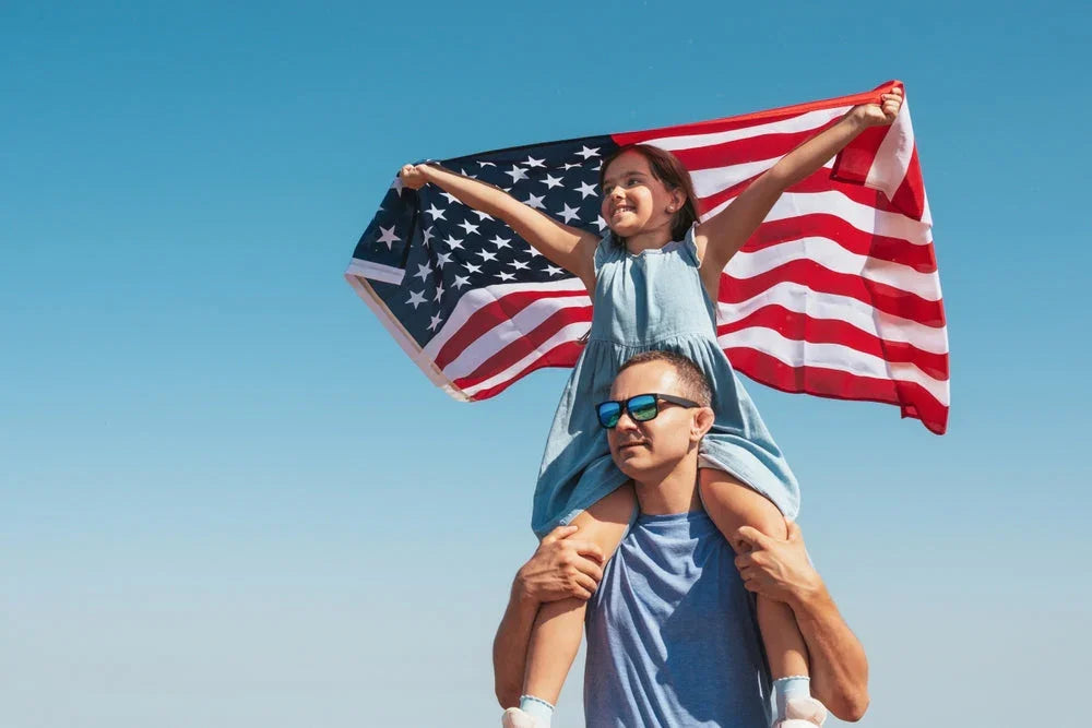 Father carrying daughter with USA flag, both in cotton clothes, under blue sky
