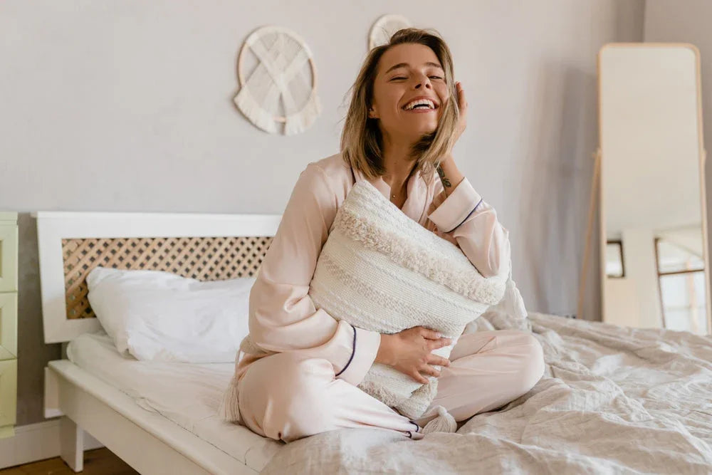 Smiling woman in soft cotton pajamas hugging pillow on bed, cozy bedroom setting