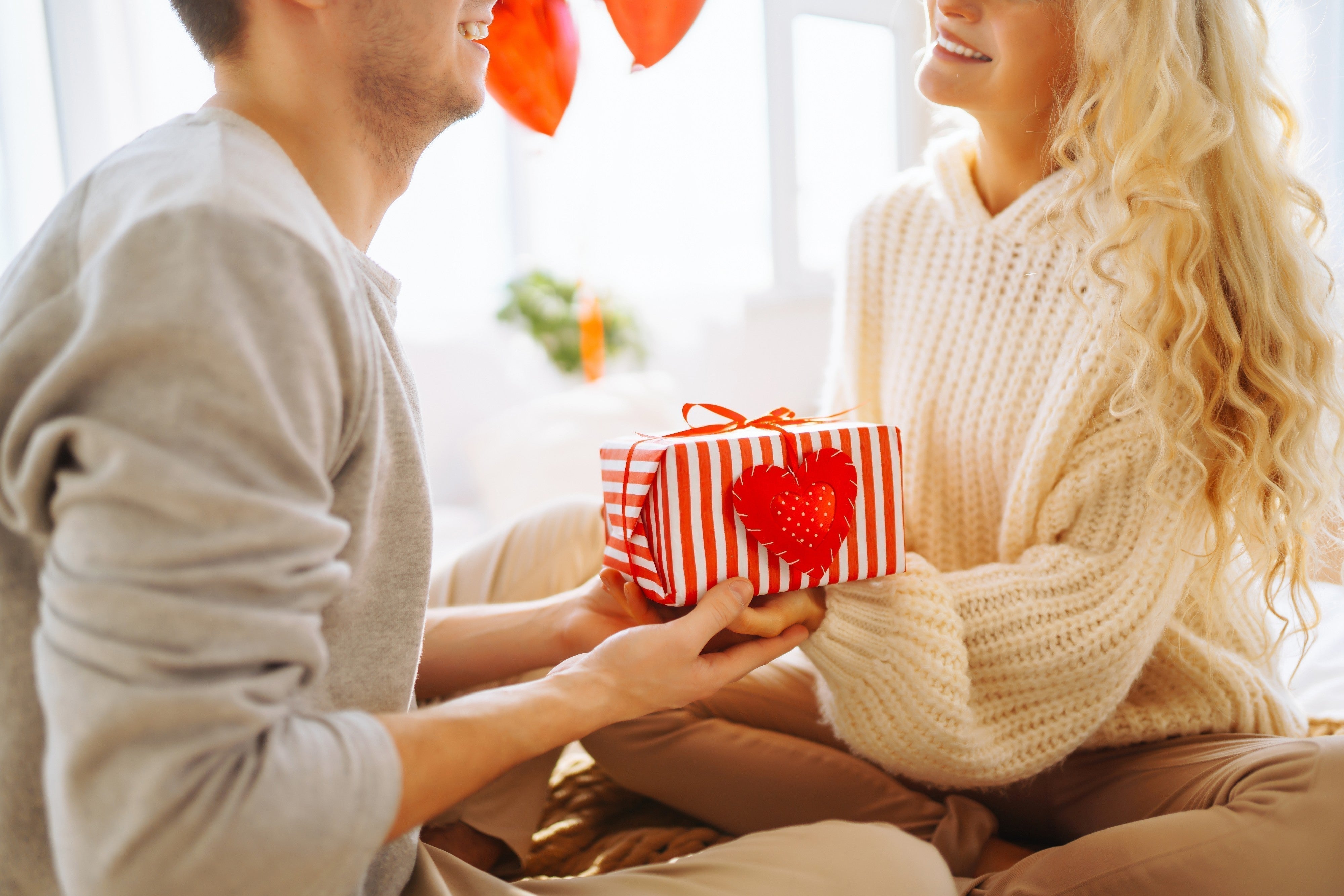 Couple exchanging gift box with heart design, wearing cozy knit and cotton clothing indoors