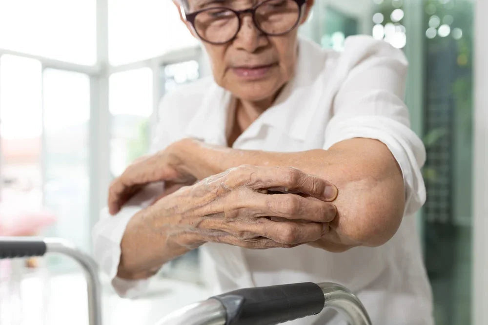 Senior woman scratching her arm, showing skin irritation, indoors with natural light.