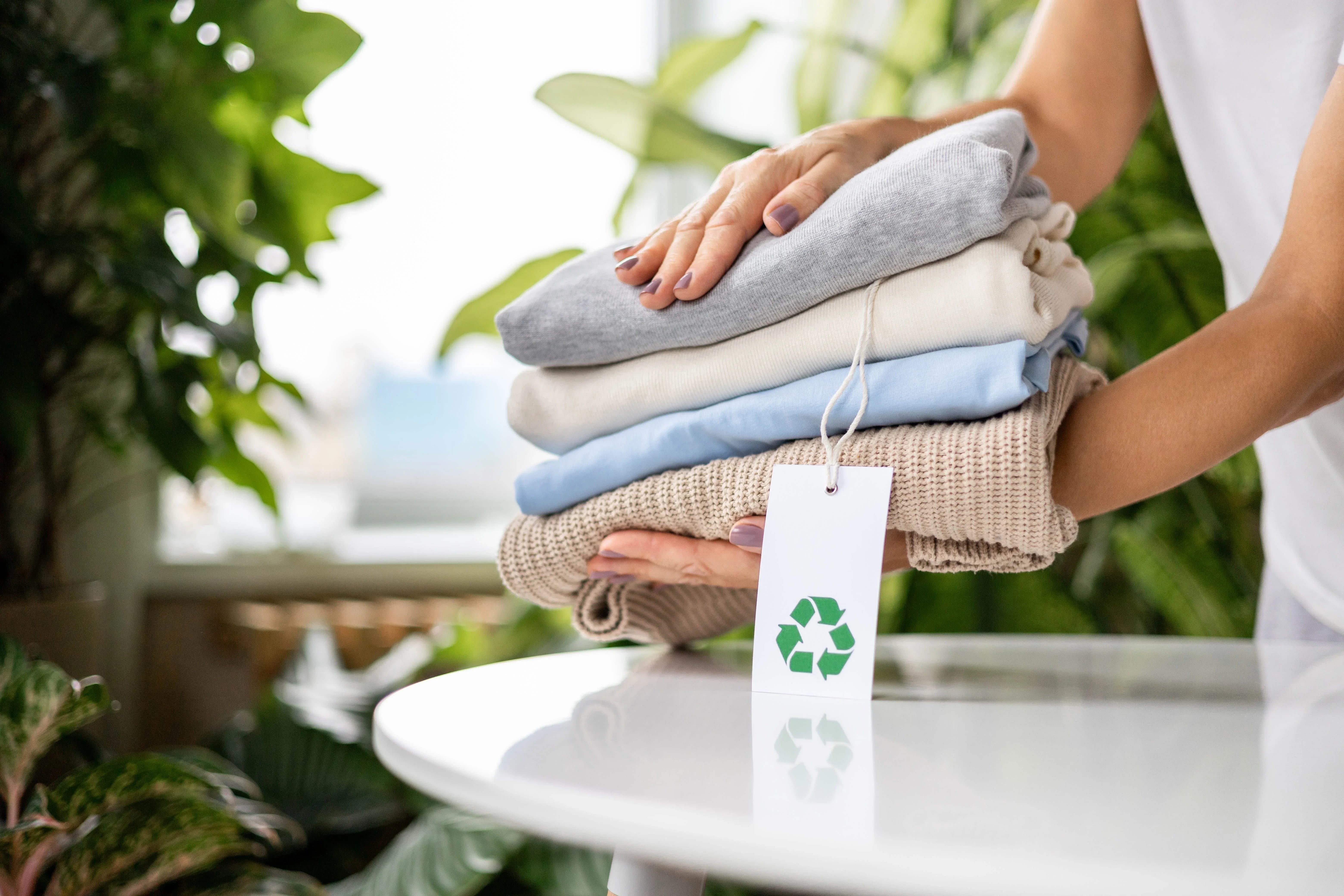 Hands holding folded hypoallergenic organic clothing with a recycling tag, surrounded by plants