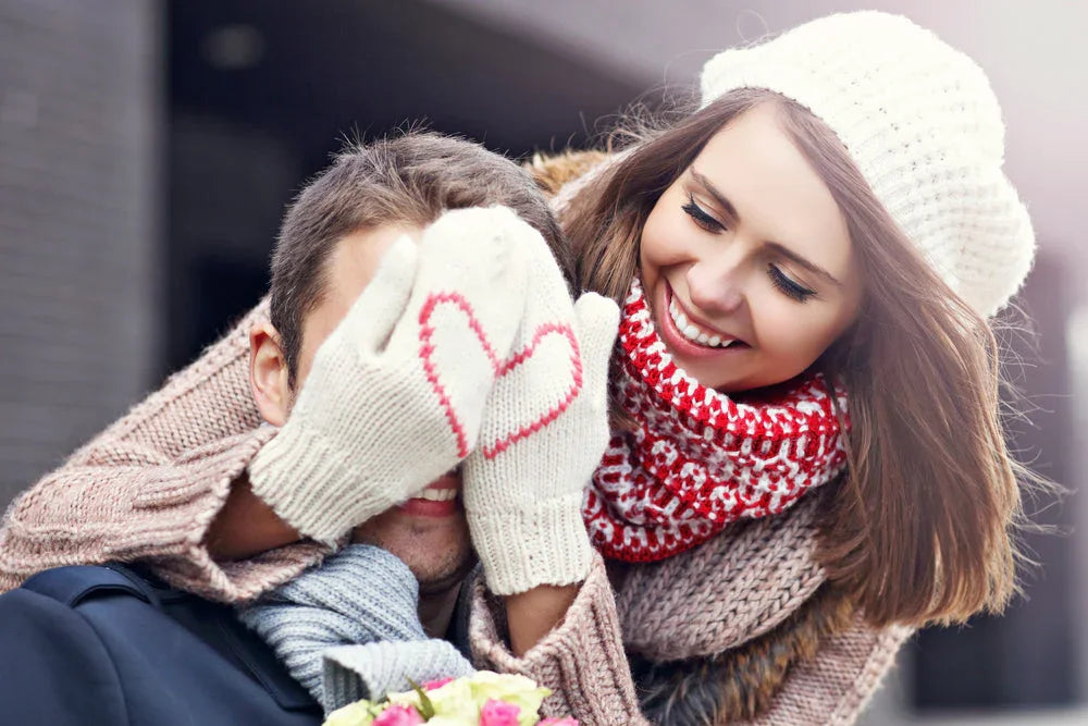 Smiling woman in allergy-free knit hat and scarf covers man's eyes with wool gloves featuring red heart.