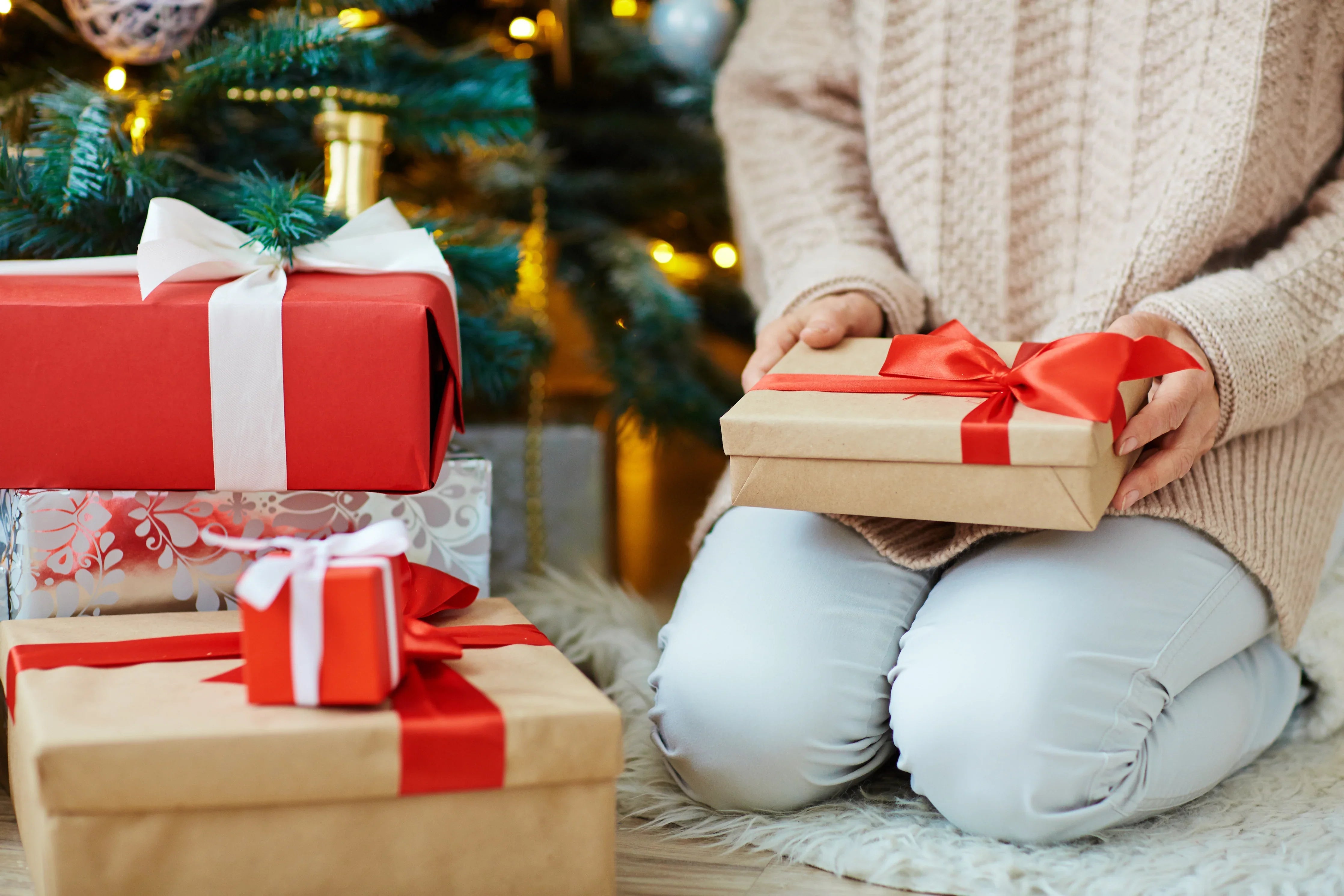 Person in cozy knit sitting by Christmas tree with allergy-free gifts wrapped in red ribbon