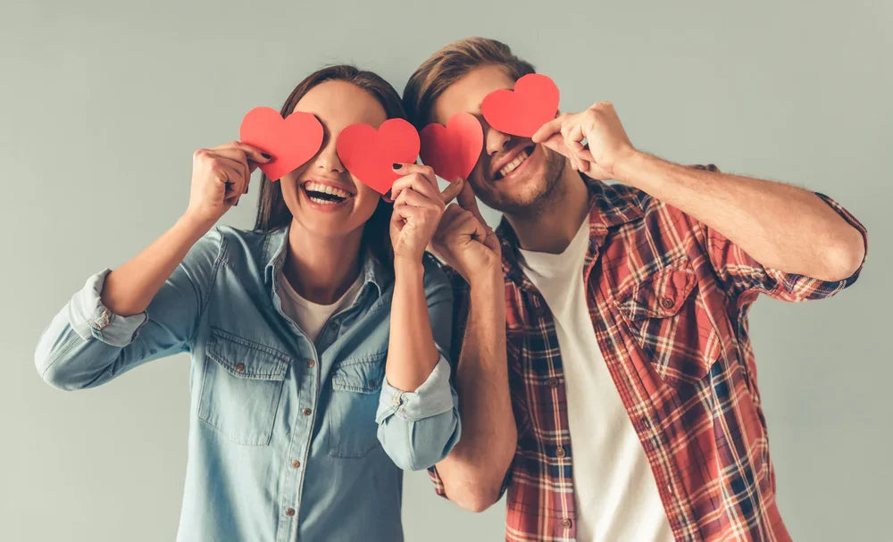Smiling couple in casual cotton shirts holding red paper hearts over their eyes
