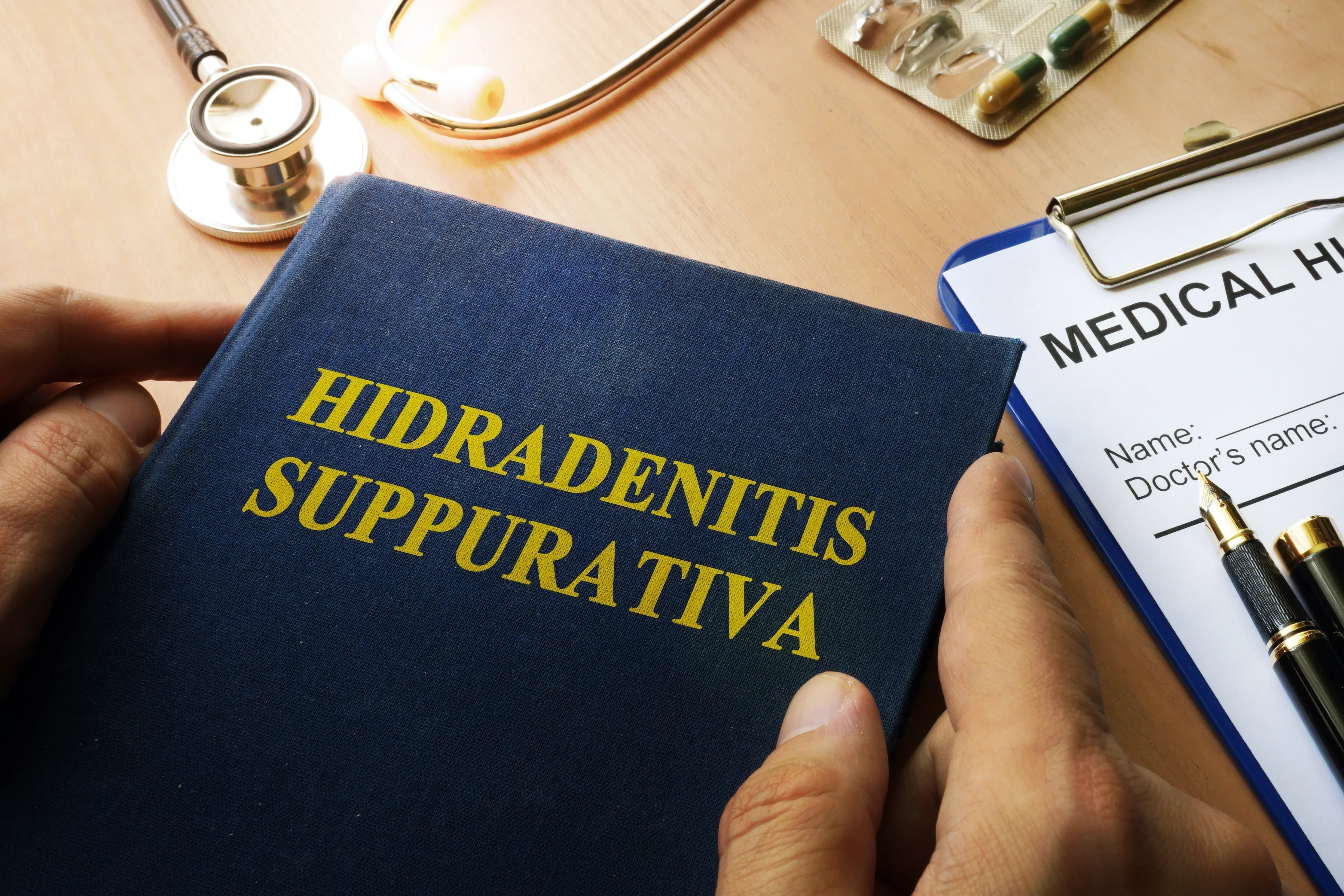 Person holding book titled Hidradenitis Suppurativa beside stethoscope and medical form