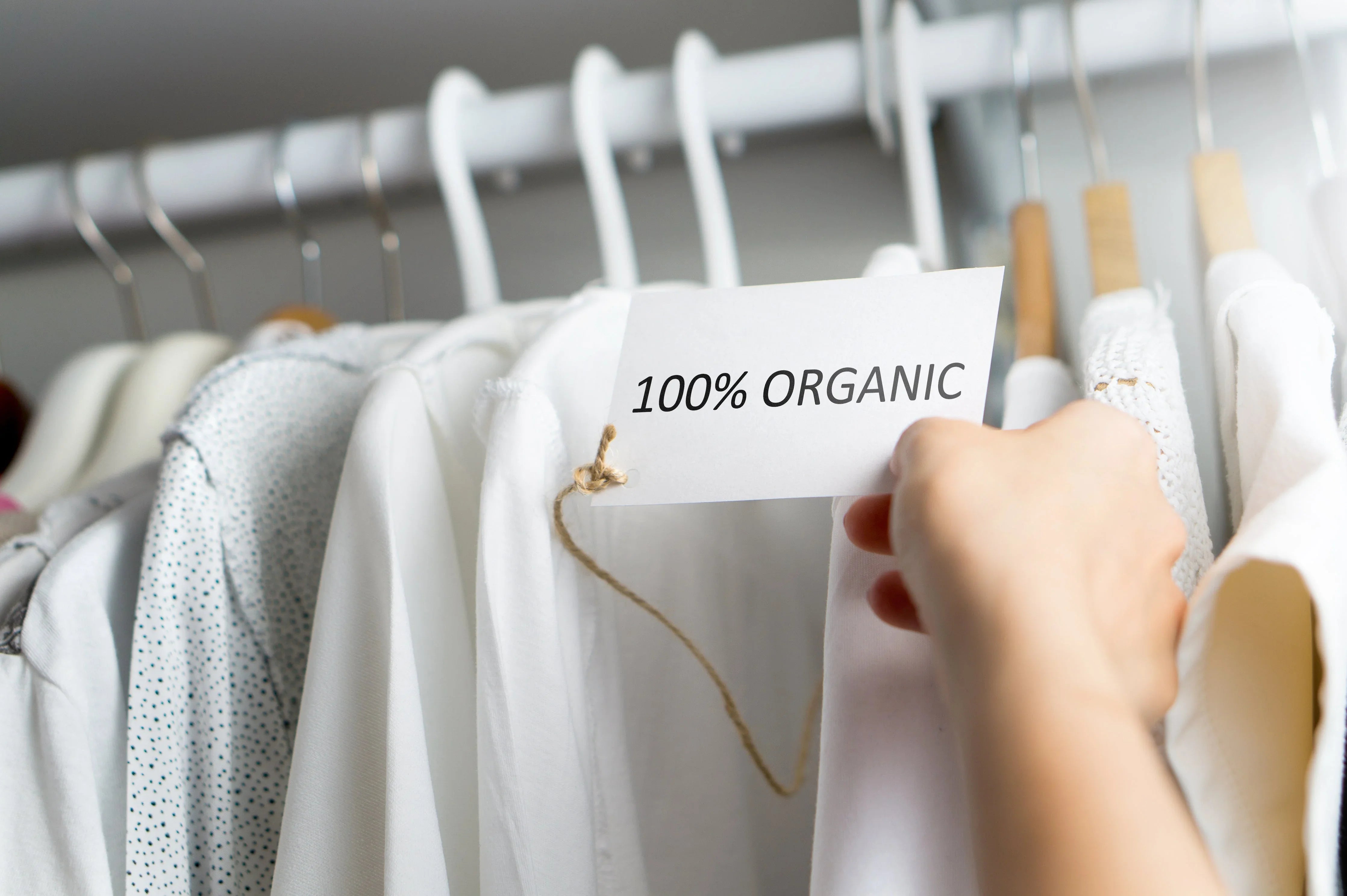 Hand holding 100% organic tag on white cotton clothes hanging in a closet
