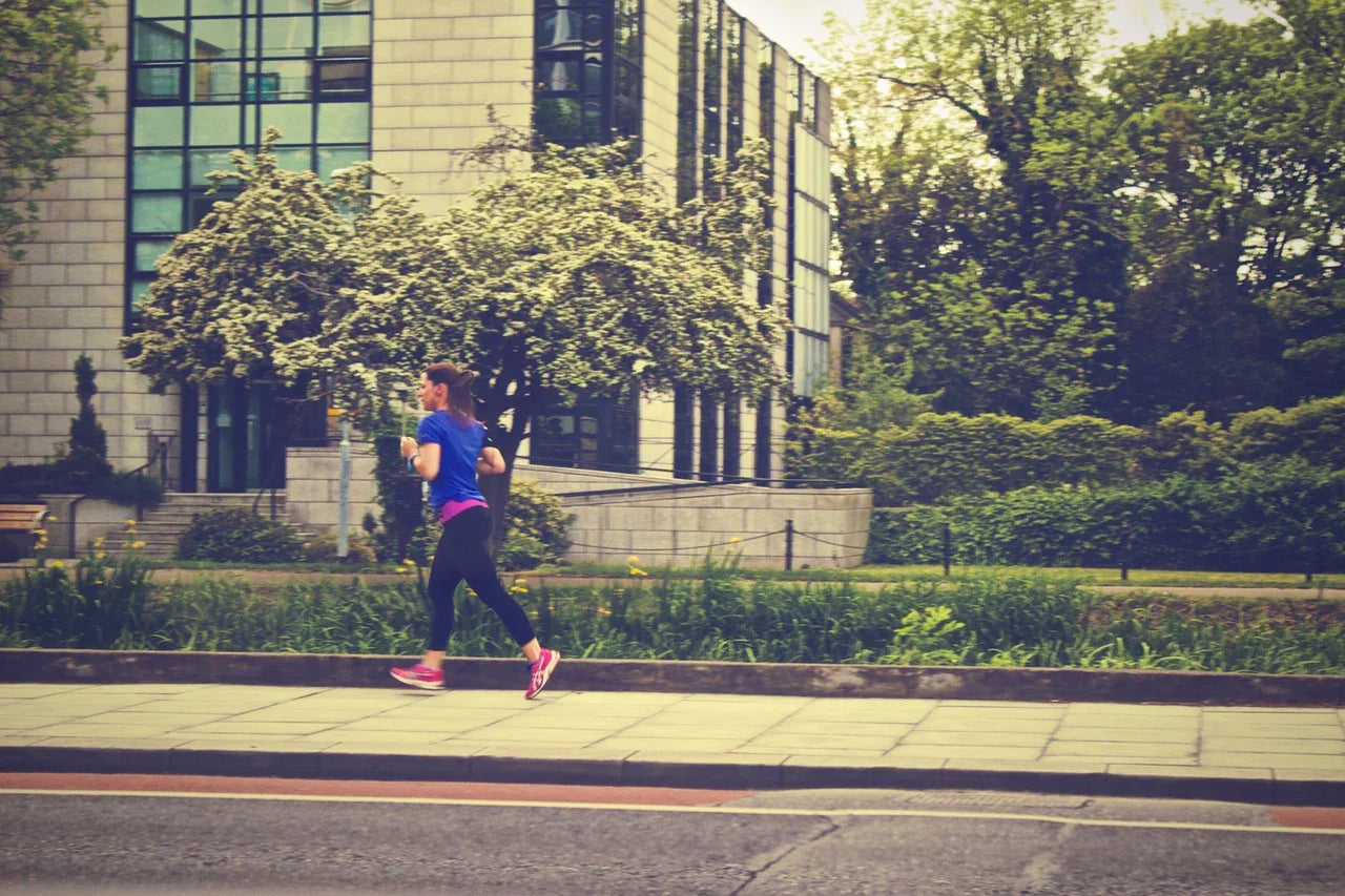 Woman jogging outdoors in athletic wear on a city sidewalk with greenery and modern buildings