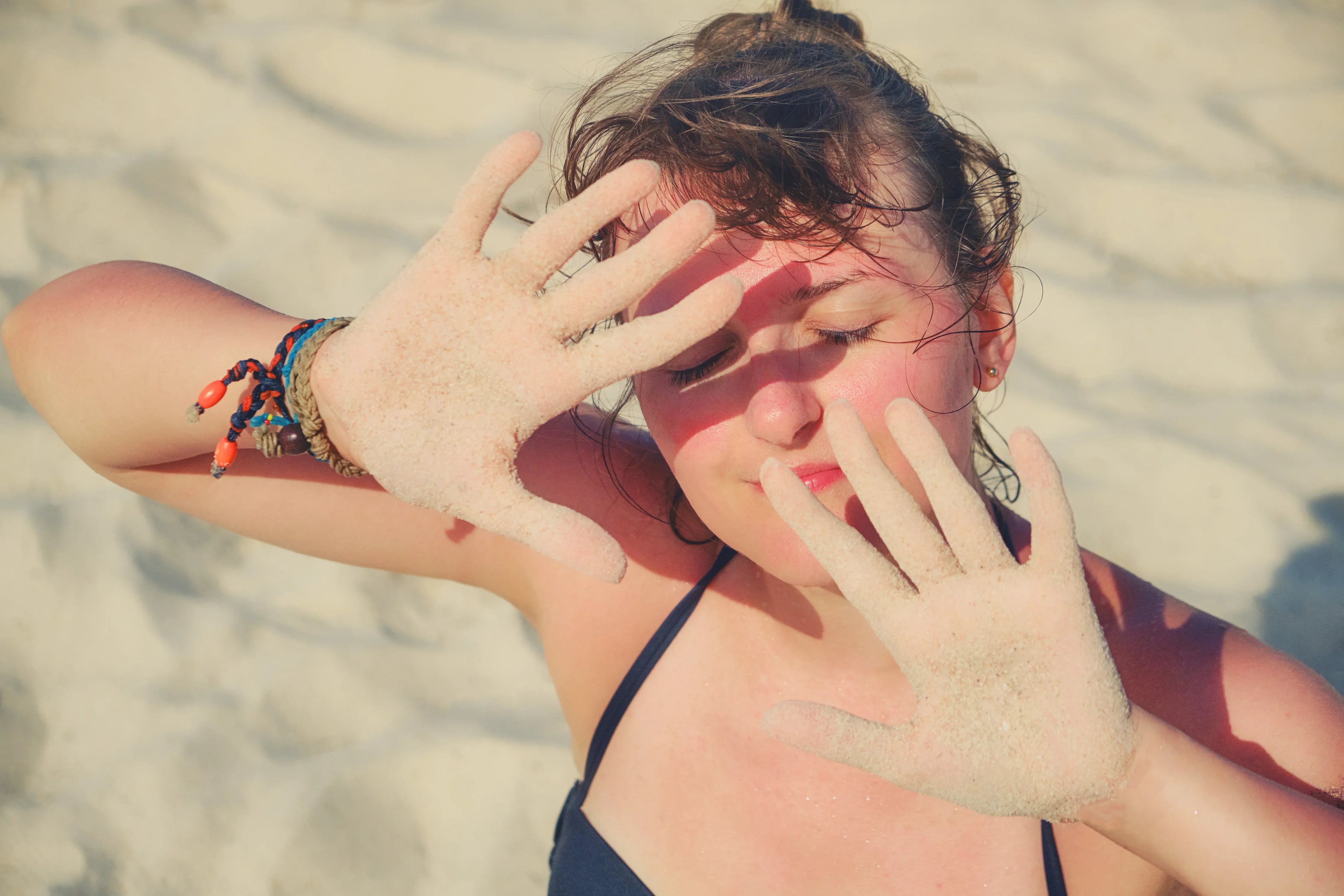 Woman in swimsuit shielding face from sun with sandy hands on a bright beach