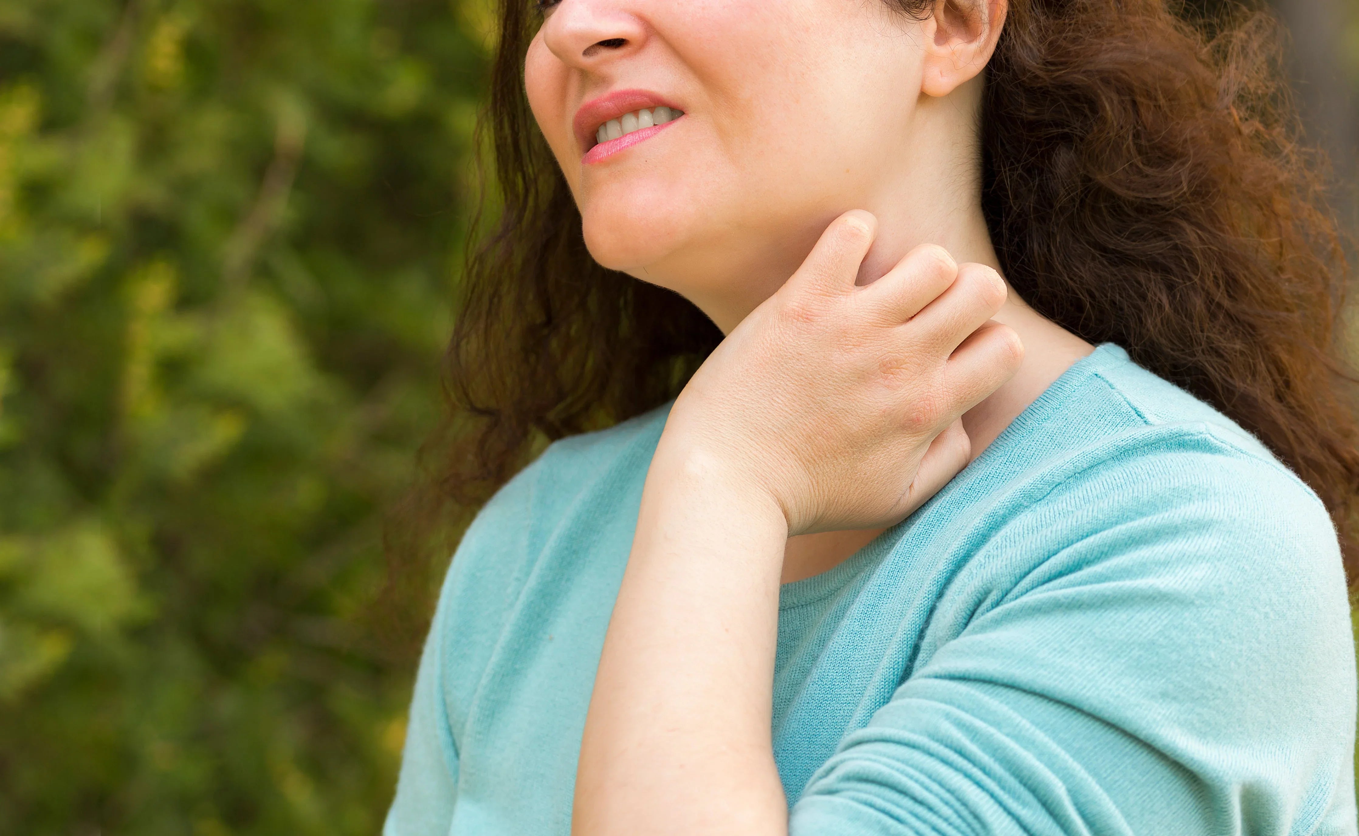 Woman in light blue shirt scratching her neck, showing skin irritation outdoors, allergy concept