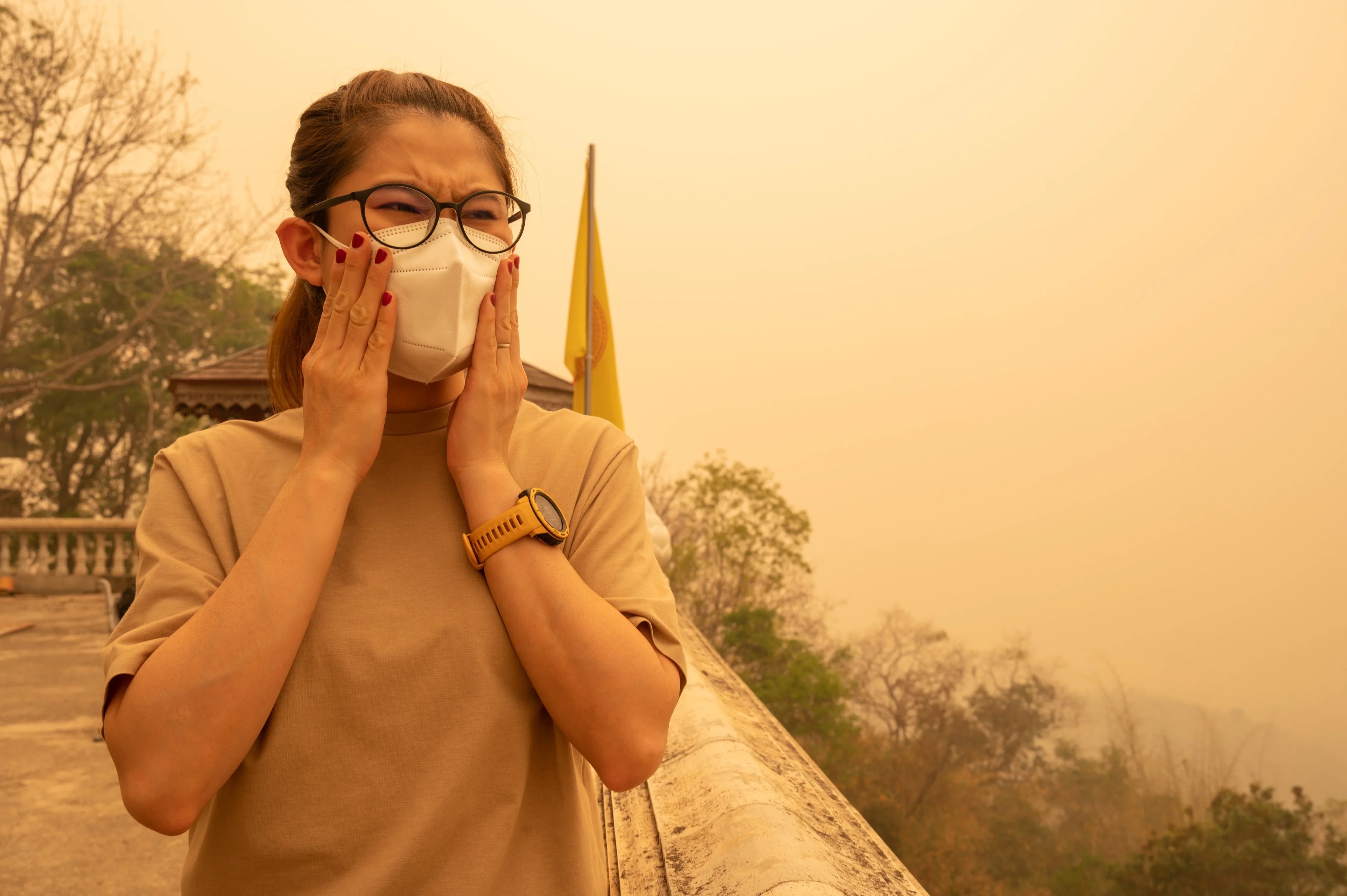 Woman wearing face mask outdoors in smoky air, touching face, trees in background